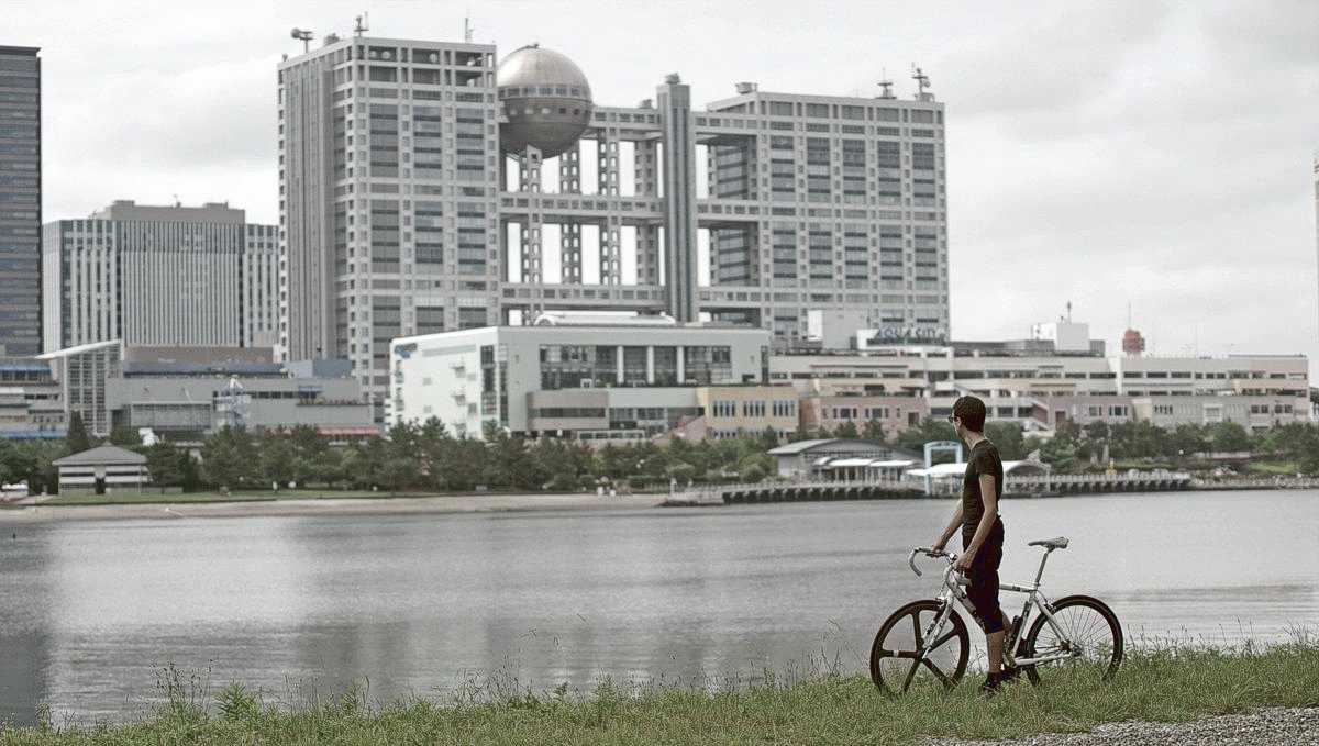 Devine in front of Fuji Terebi in Odaiba, JP Devine is on a bike on a grassy shore, facing back to us in front of water with an imposing building with a spherical wing in the background.