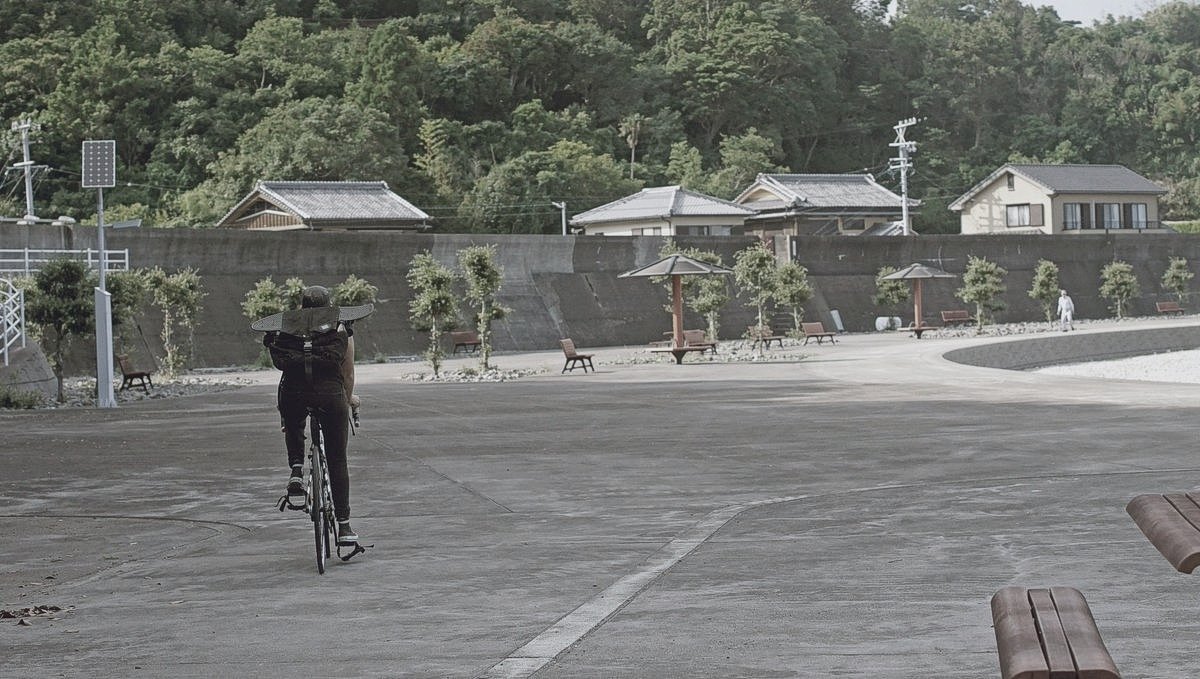 Rek cycling in Minamiise Japan Rek is cycling over concrete walkway, a skateboard strapped to their backpack, in a small Japanese ocean-side village