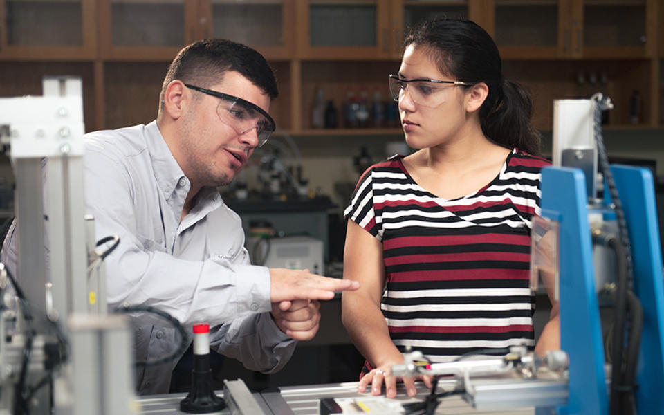 A man wearing safety glasses gestures as he explains something to a woman watching attentively, in a lab. 