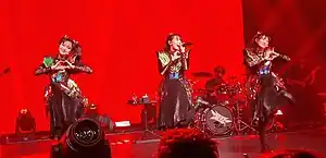 Three women wearing similar costumes consisting of a metallic top and dark skirt, performing on stage lit in bright red