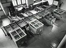 Looking down from above at a room at an intricate mechanical device which fills the room. In the background, a man sits at a desk next to a filing cabinet.Owens 1991, p. 15.