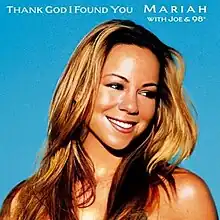 A woman with blonde hair is smiling. She wears a short white shirt and soft make-up. She crosses her right arm behind her head and is pulling a portion of her shirt with her left hand. She is standing in front of a wall with a cloud and a rainbow on it.