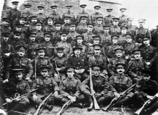 A formative photograph of soldiers in First World War British uniforms, standing and sitting in six rows. A gentleman in civilian clothes sits in the centre of the second row from the front.