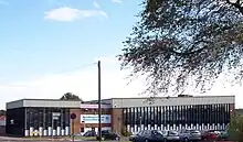 A building approximately 3 stories high. For the most part, the building is made up of horizontal columns of an opaque white panel up to around head height, with 3 windows arranged vertically above this. The columns are approximately 0.5&nbsp;m (1.6&nbsp;ft) wide and run from just below the roof to just above ground level and are separated by vertical wooden beams. Above the columns of windows a corrugated white border runs along the top of the building.